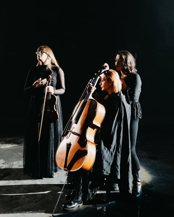 Three women in black dresses play cello and violins on a dark stage.